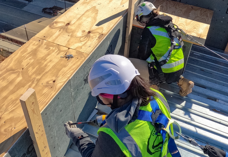 Двое строительных рабочих женского пола, оснащенные средствами защиты от падений, включая защитные шлемы, страховочные обвязки и другое оборудование.