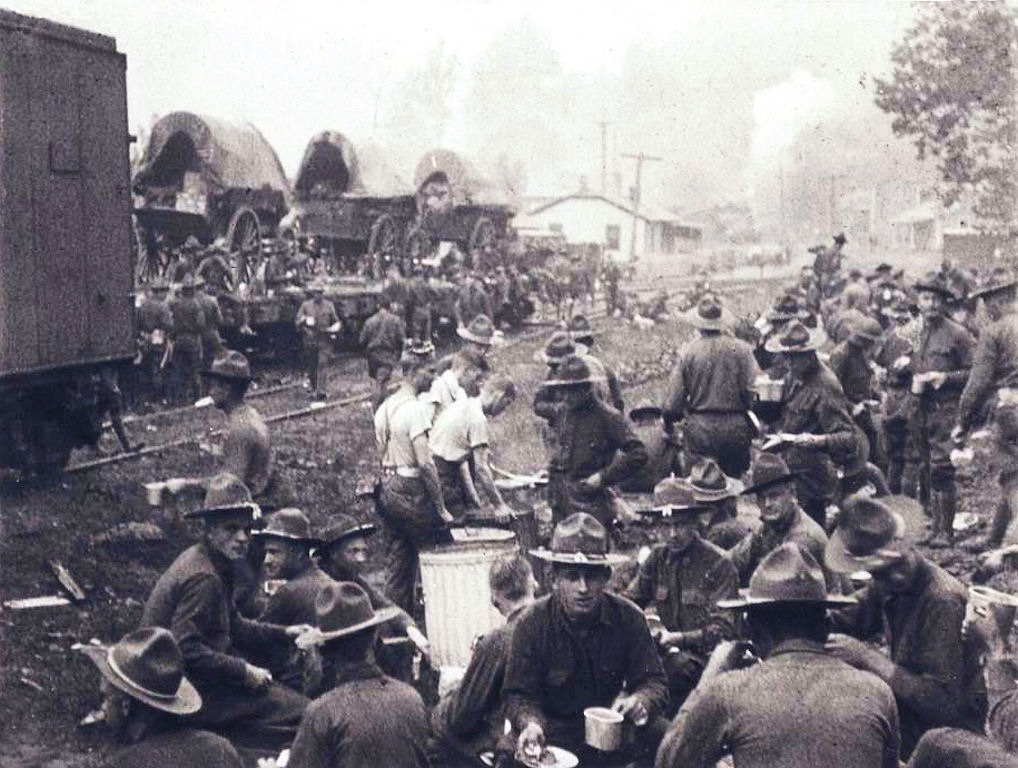 Soldiers clustered around stopped trains, many eating or drinking. Wagons with supplies can be seen on flat train cars in the background.
