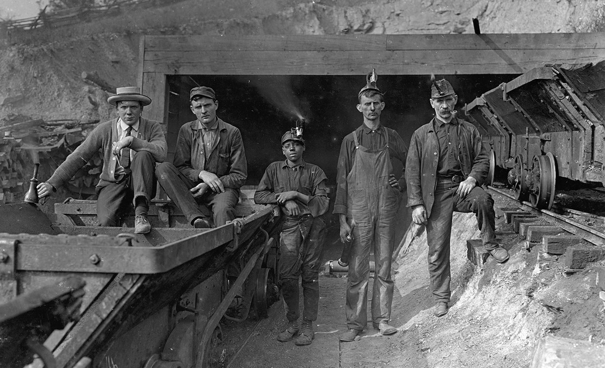 Five miners stand or sit on equipment at the entrance of a coal mine. The man in the middle is Black and the other four are white.