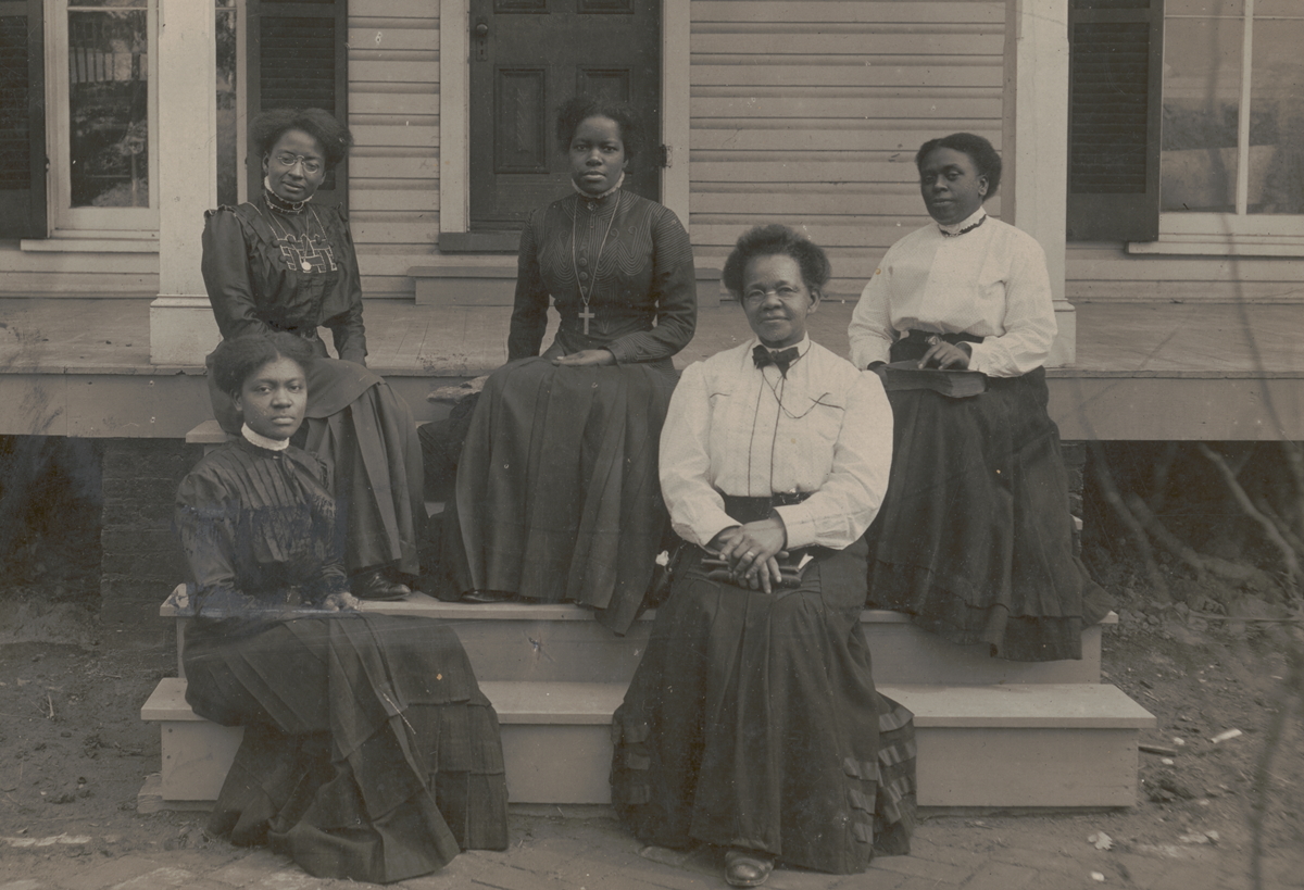 Nannie Helen Burroughs sits with four other Black women in Victorian dress on the steps to the front porch of a building.
