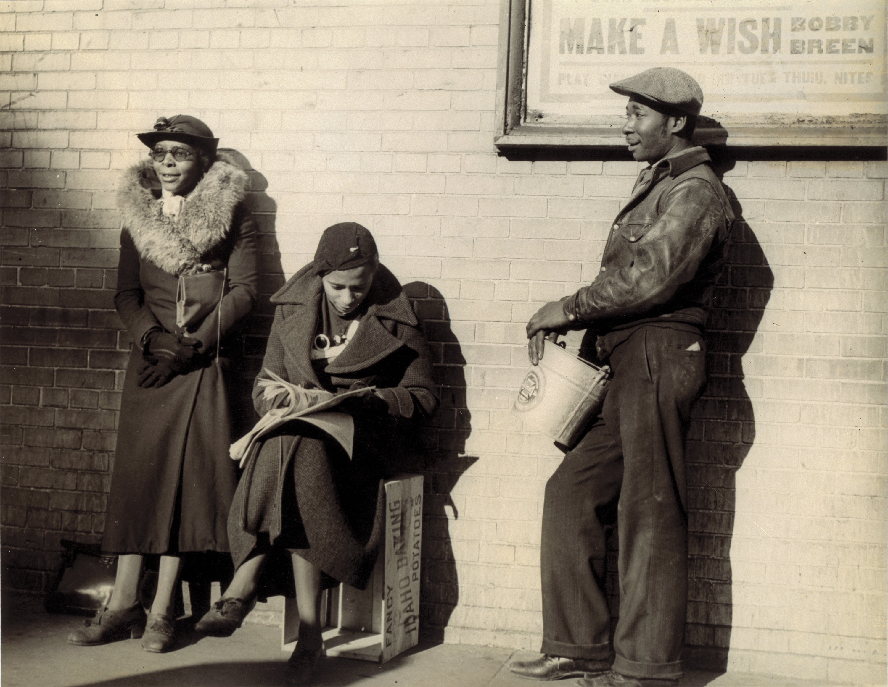 Black and white photograph showing two Black women and one man wearing coats and hats lean against a wall, waiting for employment. A movie poster behind them reads "Make a Wish."