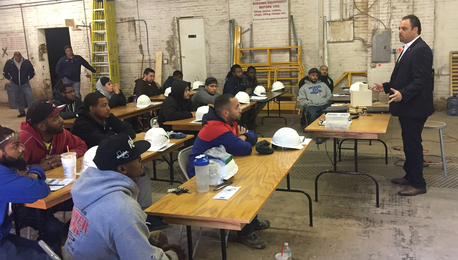A man wearing a suit addresses a room full of apprentices. They sit at folding tables with their hard hats and other work gear.