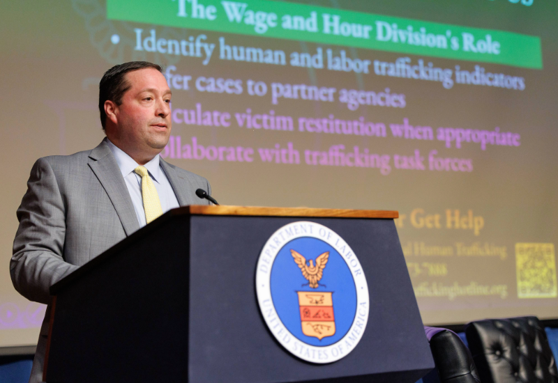 The image shows a person standing at a podium with the U.S. Department of Labor seal on it. The person is dressed in a suit and is delivering a speech. A presentation slide is visible in the background, related to human and labor trafficking topics.