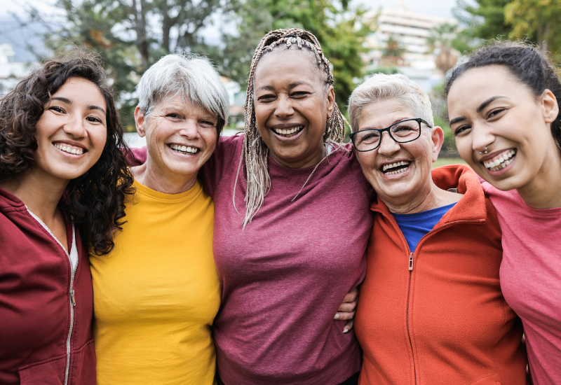 A diverse group of women of different age ranges wearing colorful shirts and jackets hugging each other and smiling for the camera.