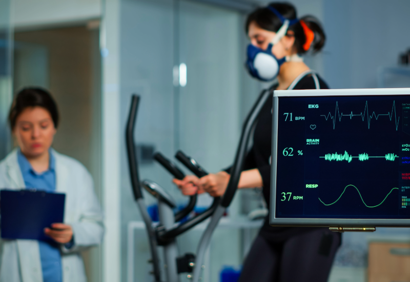 An exercise physiologist takes notes while collecting data from a woman on an elliptical machine who is wearing a respirator.