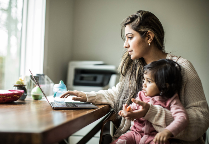 A woman types on a laptop in a home office while holding a small child. 