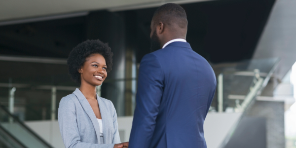 A young woman with a blue blazer smiles while shaking the hand of a man in a blue suit. They are standing in a modern office lobby. 