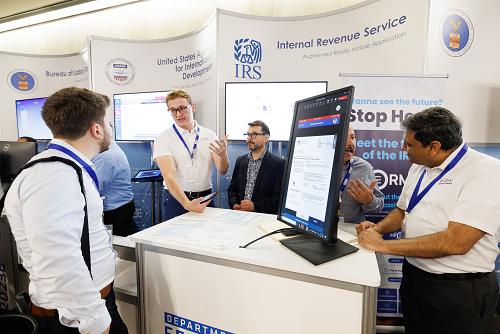 IRS representatives stand at a Tech Day booth with two large screens, speaking with event attendees.