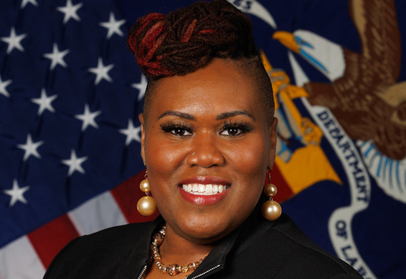 A black woman wearing professional attire smiles for a professional headshot with the American Flag serving as the backdrop. 