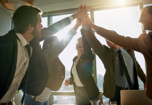 A group of professionals embracing in a high five in an office setting with the sunlight shining in through a large window.