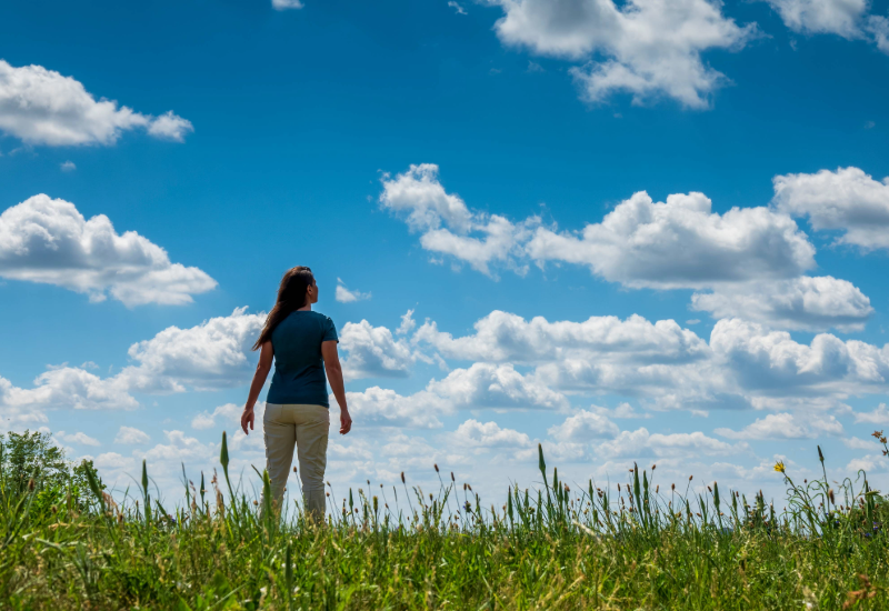 A woman wearing a blue shirt and tan pants stands in an open field and stares up at the sky.