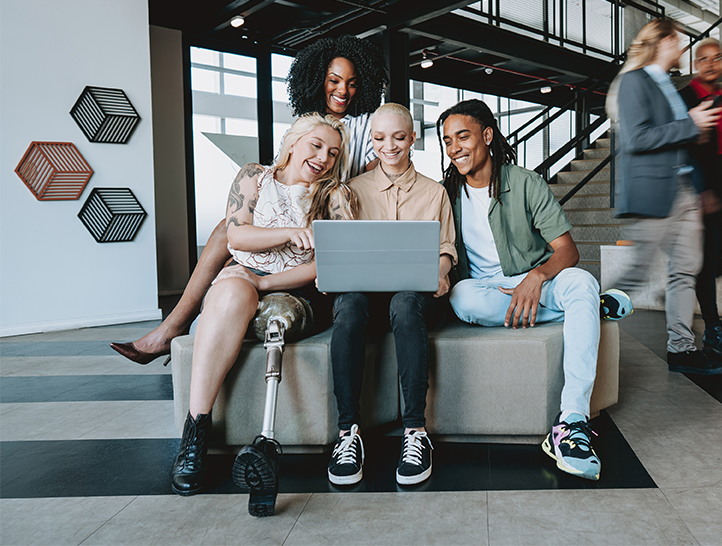 A diverse group of young people – with and without visible disabilities – sit in a building lobby, smiling as they look at a laptop screen.