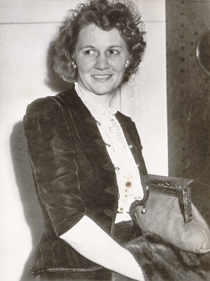 Sepia-toned photo of Elizabeth Hayes carrying a coat and purse. She has short, curly hair and is wearing a decorative jacket over a collared blouse.
