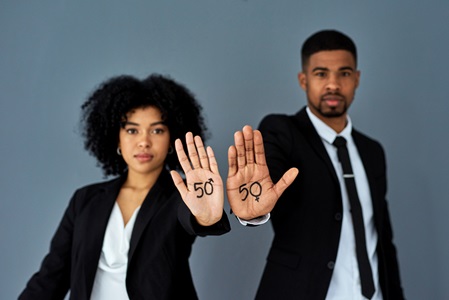 A man and woman in business suits stand side by side, holding out their hands. On both palms is written the number 50, but the 0 on the woman's hand is the male gender symbol, and the zero on the man's hand is the female symbol.