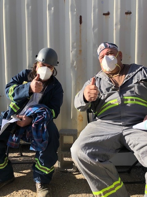 Two miners sitting on chairs, wearing masks, with their thumbs up. 