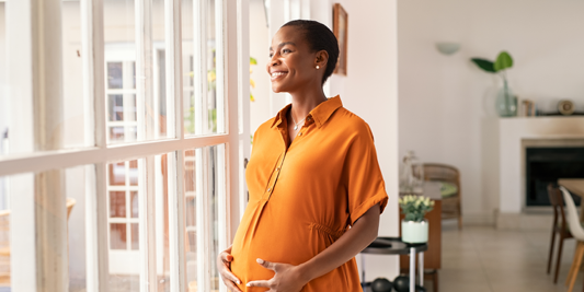 A pregnant woman smiles with her hands on her stomach while looking out a window.