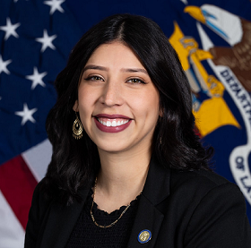 Official portrait of Nezly Silva. She has long, dark hair, and is seated in front of the U.S. and Labor Department flags.