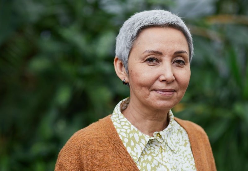 An older woman with short grey hair wearing a brown sweater and button down shirt.