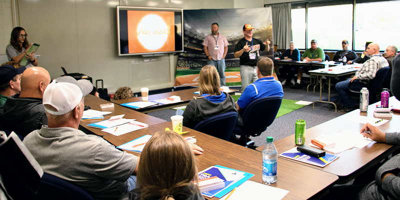 A group of safety trainers sit around tables in a classroom writing in notepads while they listen to a speaker. A screen behind the speaker has the image of a baseball with the words 