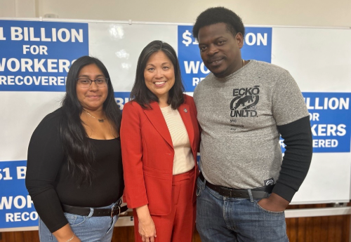 A woman with long dark hair wearing a black shirt and jeans stands with a woman with collar-length black hair wearing a red suit and a man with short dark hair wearing a grey t-shirt and jeans. They are smiling for a photo and standing in front of a white board with signs that read, " $1 BILLION FOR WORKERS RECOVERED."