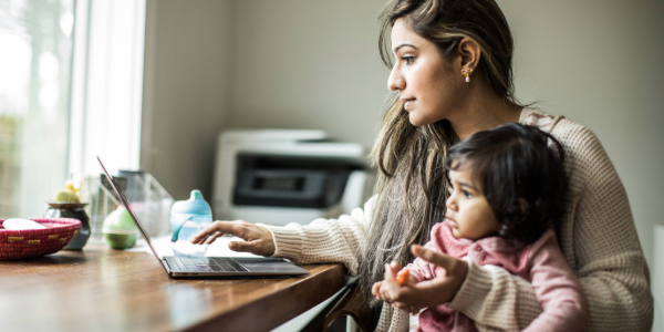 A woman types on a laptop in a home office while holding a small child.