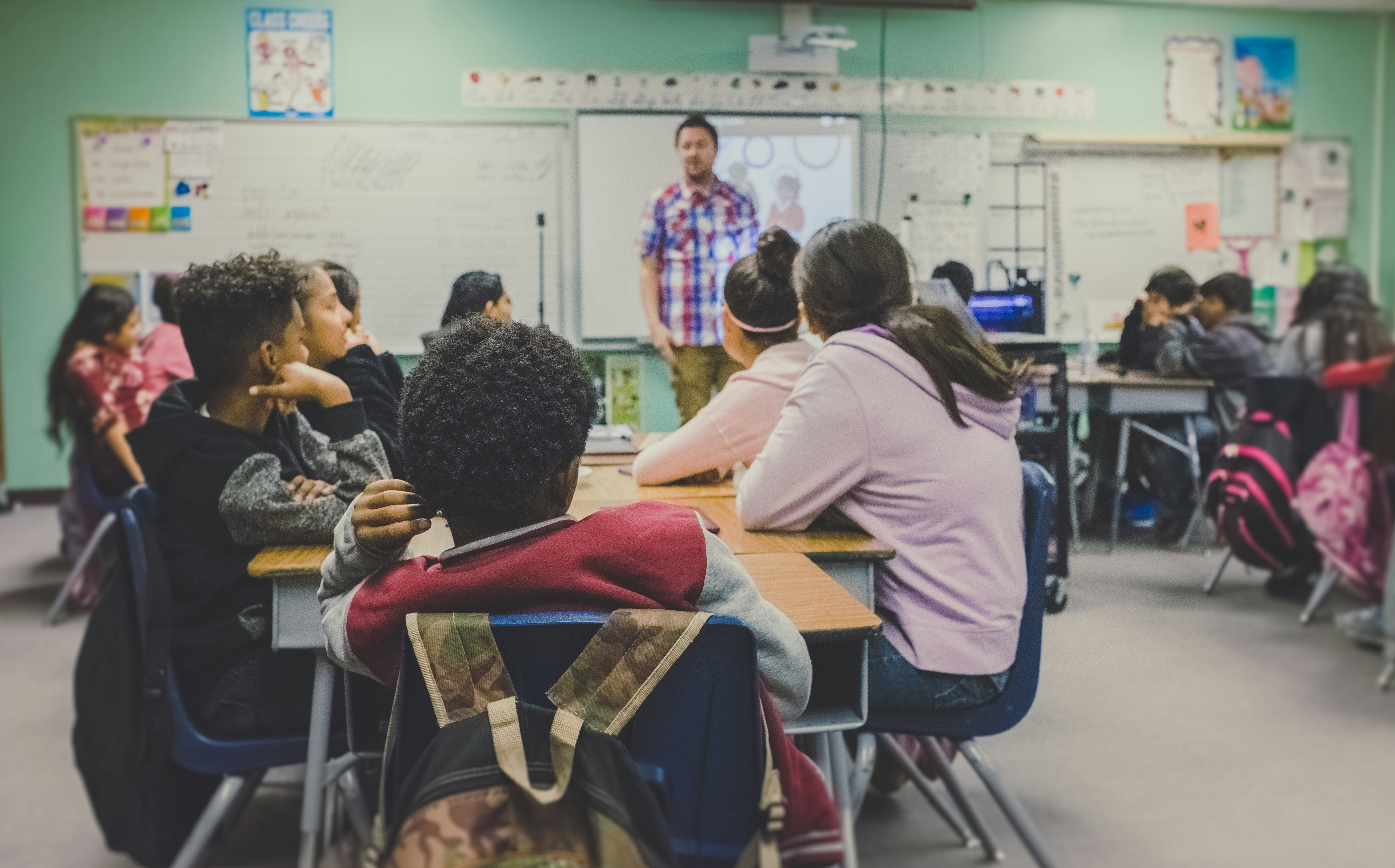 A teacher stands in a classroom addressing a dozen teenagers seated at desks with backpacks on the chairs.