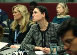 A woman with dark hair pulled back and wearing a black and white jacket listens intently during a panel on global migration.