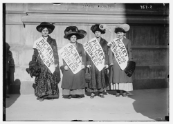Black and white photo of garment workers striking during the "Uprising of the 20,000" in New York City, February 1910. Four women wearing long layered dresses and large hats are also wearing sashes with "Picket Ladies Tailors Strikers"