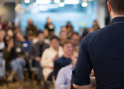 A stock photo showing the back of a male speaker as he addresses an audience 