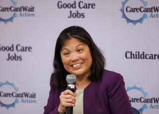 Acting Secretary Julie Su holding a microphone in front of a backdrop that says Good Care Jobs and Care Can't Wait Action