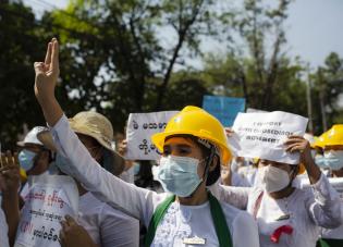 Workers wearing white clothing and face masks protest in Myanmar. Many are holding signs. Credit: AHS