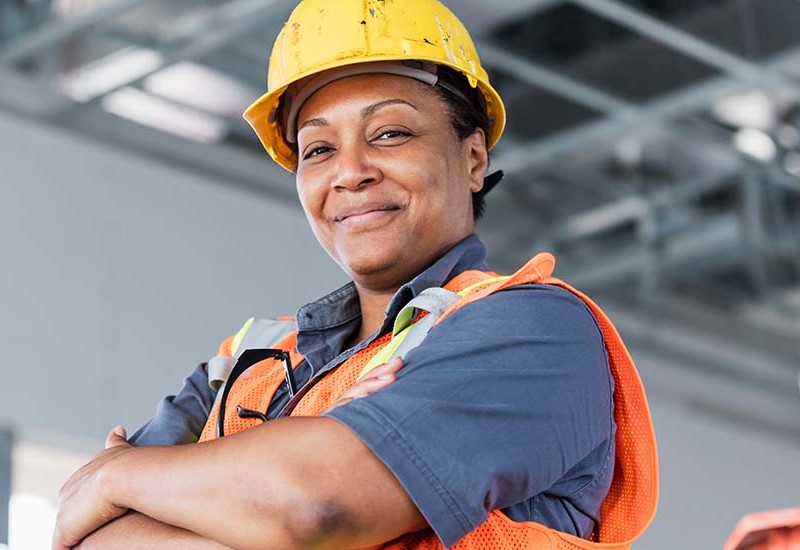 A Black woman wearing protective equipment and a hard hat. She is smiling for a photo with her arms folded.