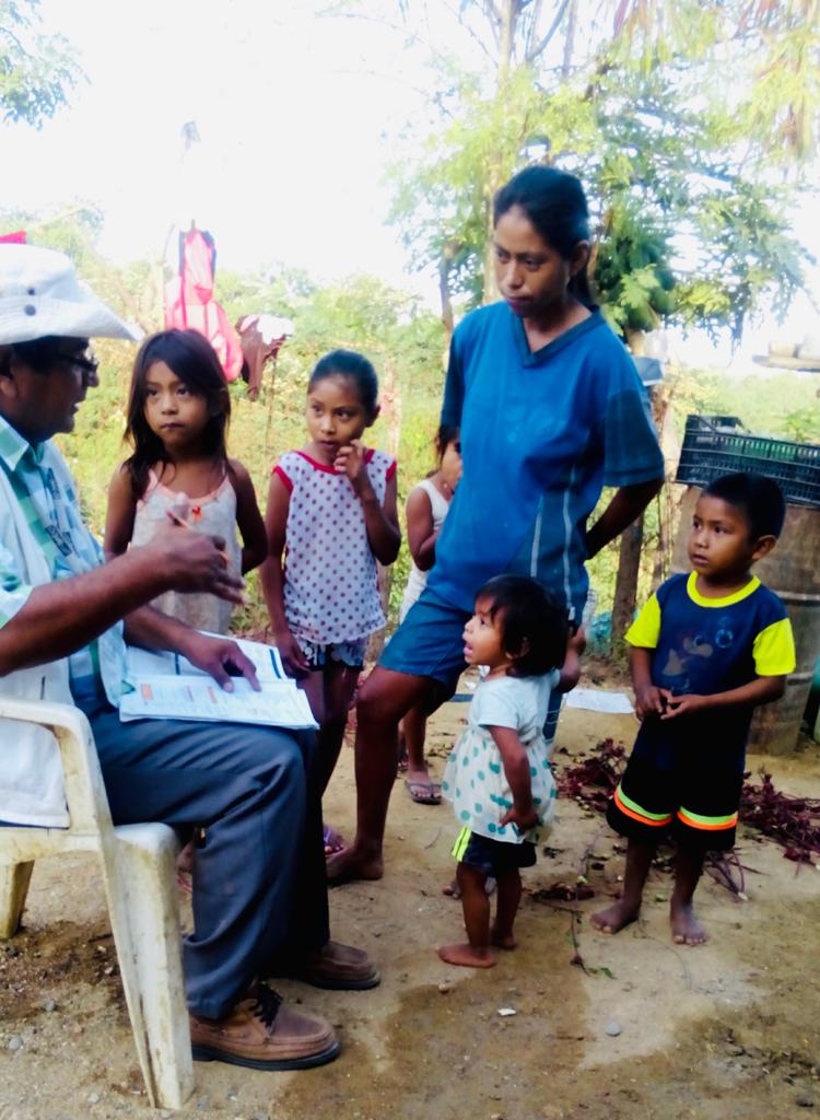 A man holding paperwork and a pencil speaks outdoors with a woman who is surrounded by several small children.