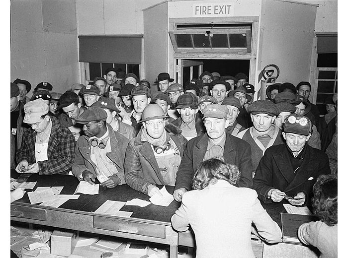 Black and white 1940s photo showing a crowd of men in work clothes clustered inside an office. There are so many that they are spilling out the door.