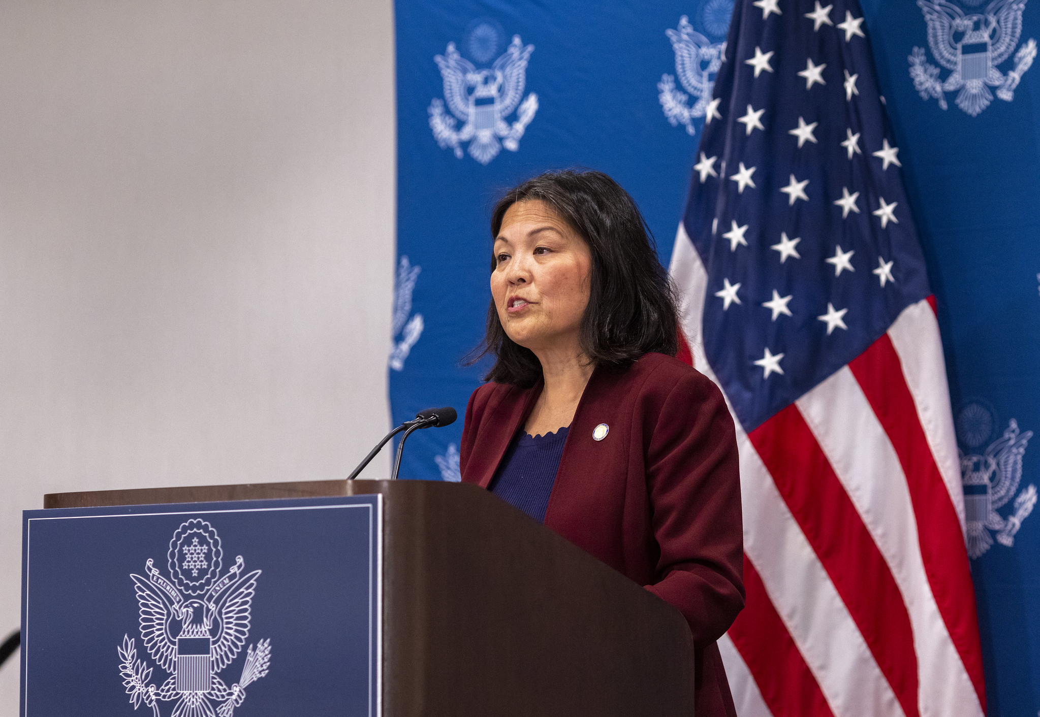 Julie Su standing at a podium with the U.S. flag behind her.