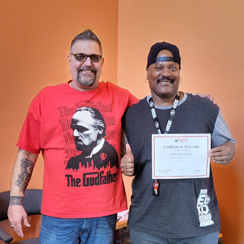 Two men stand side-by-side, smiling for the camera. The man on the left wears glasses and a Godfather tee-shirt. His arm is around the other man, who wears a 49ers lanyard and holds a Certificate of Training. He is giving the thumbs up signal. 