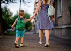 A rear view of a mom wearing a blue dress and flat shoes walking with her son wearing a grey shirt, blue shorts, sneakers and a green backpack while holding hands.