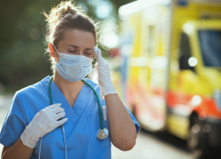 A stressed woman in scrubs with a stethoscope and medical mask outdoors near a yellow ambulance.