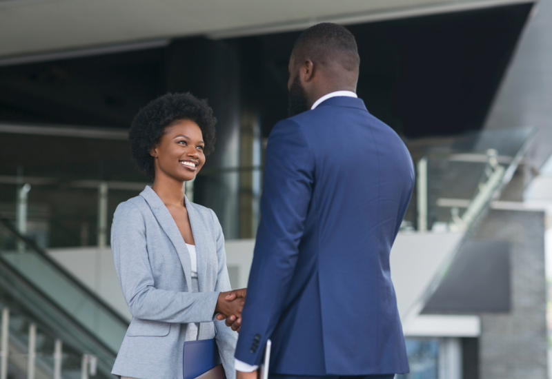 A young woman with a blue blazer smiles while shaking the hand of a man in a blue suit. They are standing in a modern office lobby. 