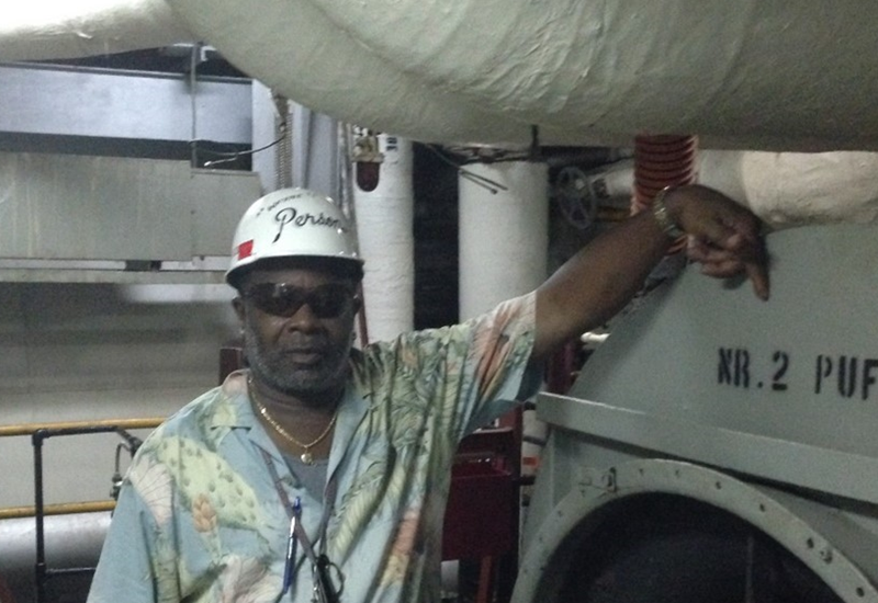 A man stands on a ship wearing a hardhat and protective sunglasses. 