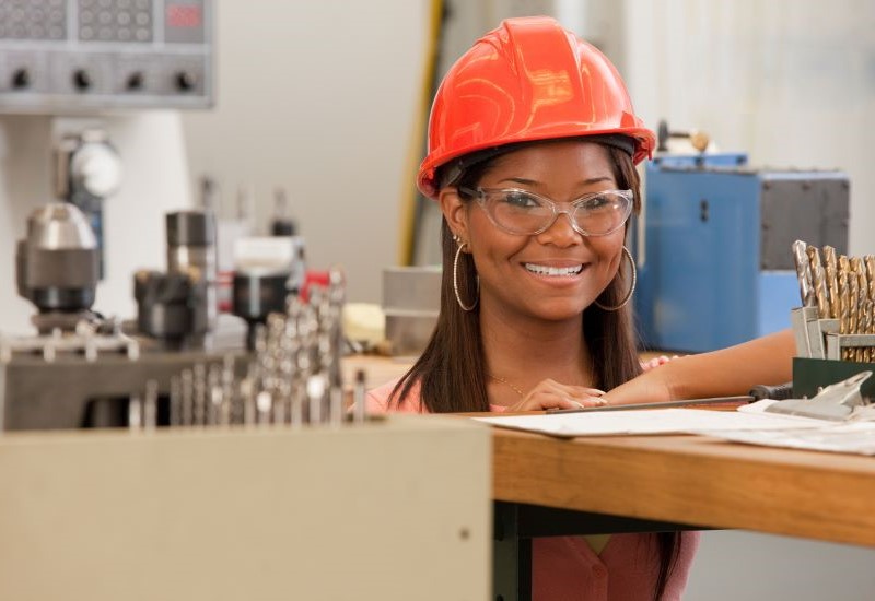 A young woman in a hardhat and safety goggles smiles while standing in a workshop.