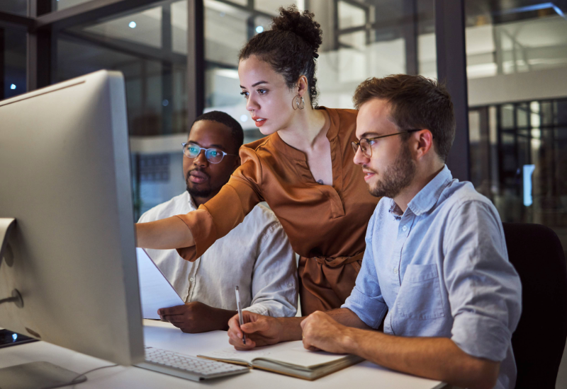 Three workers lean over a desk, looking at a computer screen.