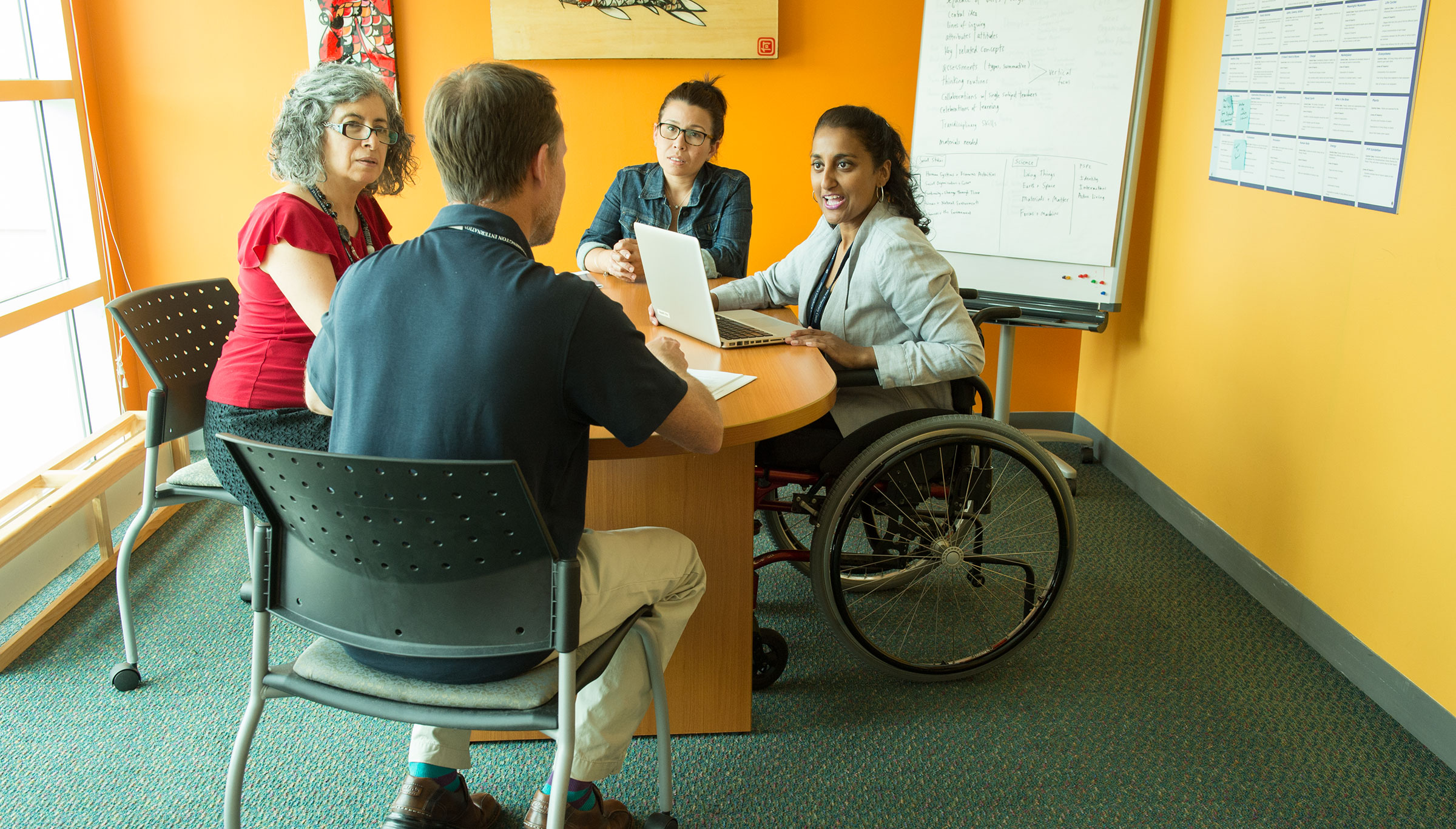 A photo of an inclusive workplace, with four men and women seated around a table in a conference room. One of the women is using a wheelchair.