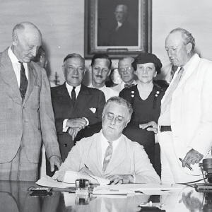 Black and white photo: President Franklin Roosevelt signs the Social Security Act, flanked by supporters, including Secretary Frances Perkins.