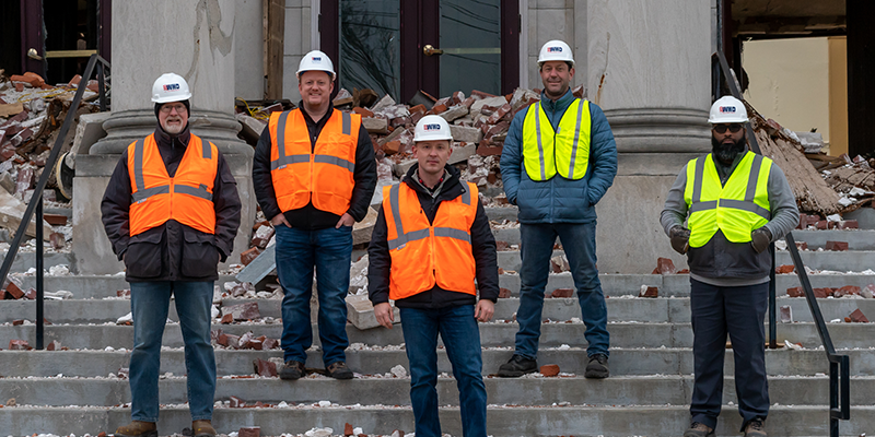 Five men in safety vests and hard hats stand on the crumbling steps of a storm-damaged building