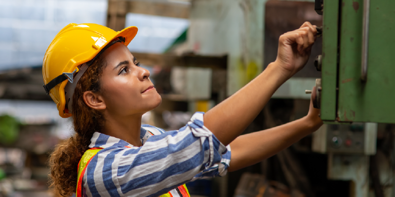 A woman wearing a hard hat works in a machine shop or industrial environment