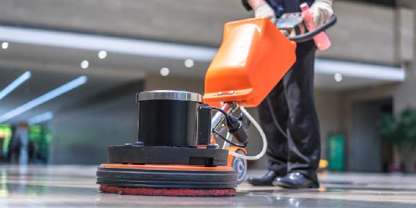 A custodian uses a floor waxer in a building lobby.