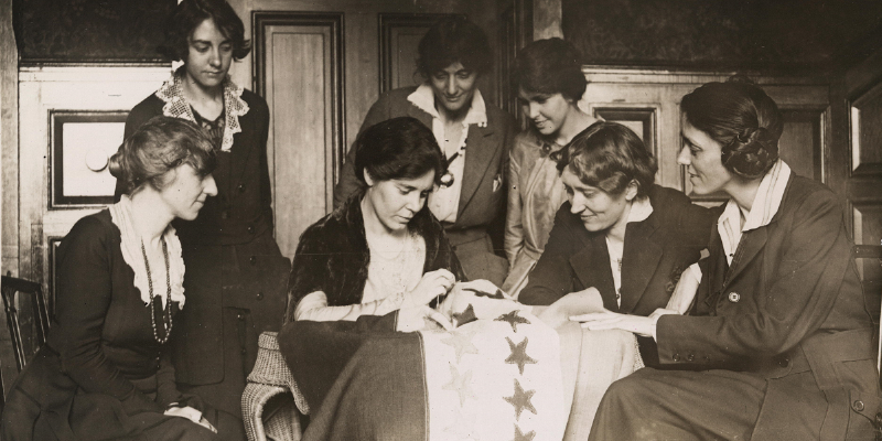 Black and white photo shows seven women sewing a flag in 1920.