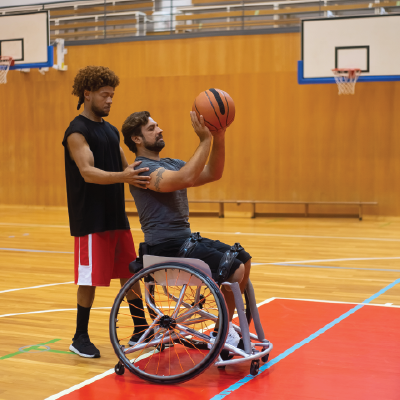 A personal trainer guides a man in a wheelchair as he prepares to shoot a basketball. Photo by Kampus Production.