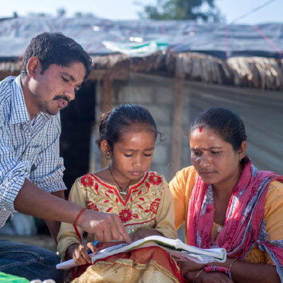 A father and mother read with their daughter in front of their home in Nepal. 